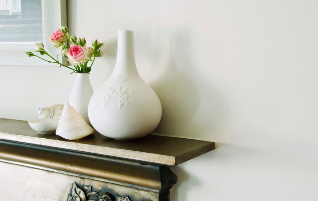 Add some greenery. Two white vases with pink roses in them on the fireplace mantel.